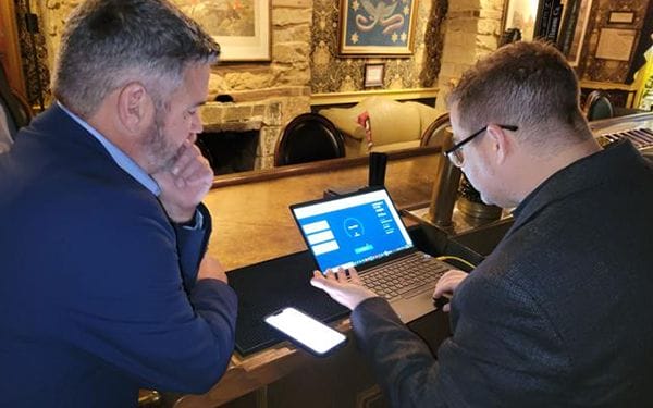 Two men in suits stand at a bar counter, one typing on a laptop displaying a blue screen with charts and text, while the other observes thoughtfully. A smartphone lies on the counter beside them.