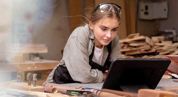 A young woman wearing safety glasses and an apron leans over a workbench in a woodworking shop, looking at a tablet. Various woodworking tools and wooden pieces are scattered on the table around her.