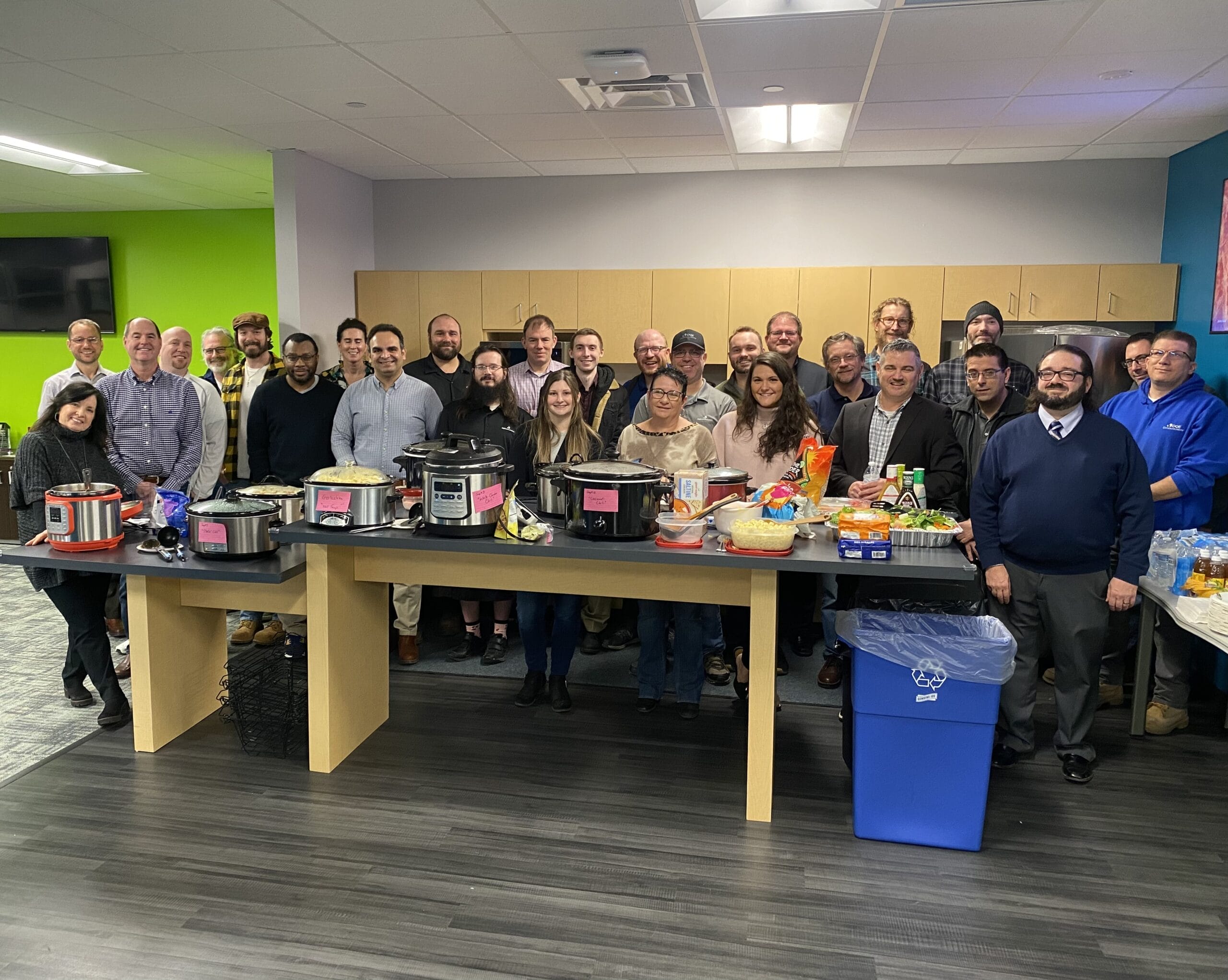 A group of about 25 adults poses and smiles behind a long table filled with slow cookers, food, and snacks in an office breakroom with bright green and blue walls.