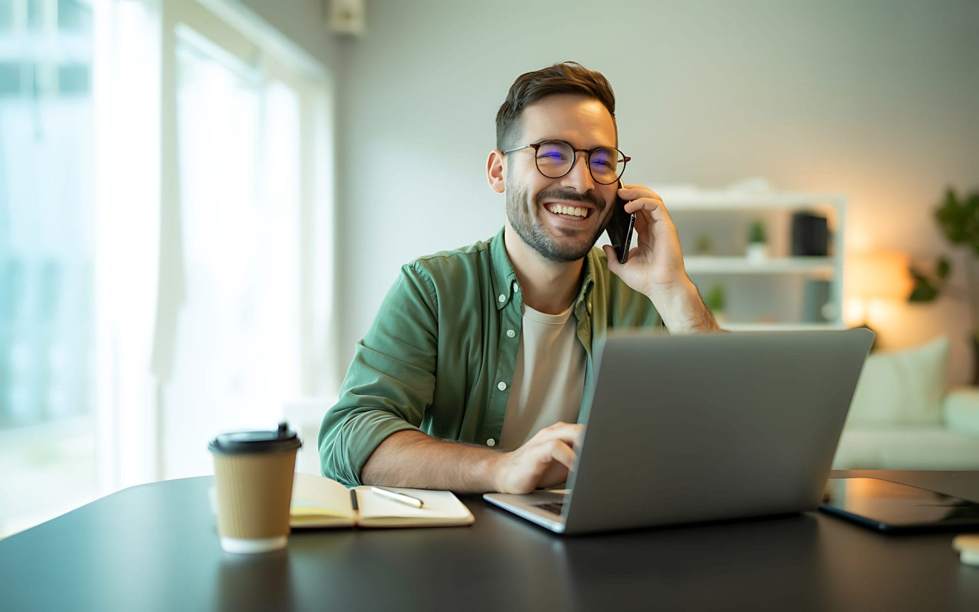 A smiling man with glasses talks on the phone while using a laptop at a desk. There is a notebook, pen, and takeaway coffee cup beside him. The background shows a bright, modern room with shelves and plants.