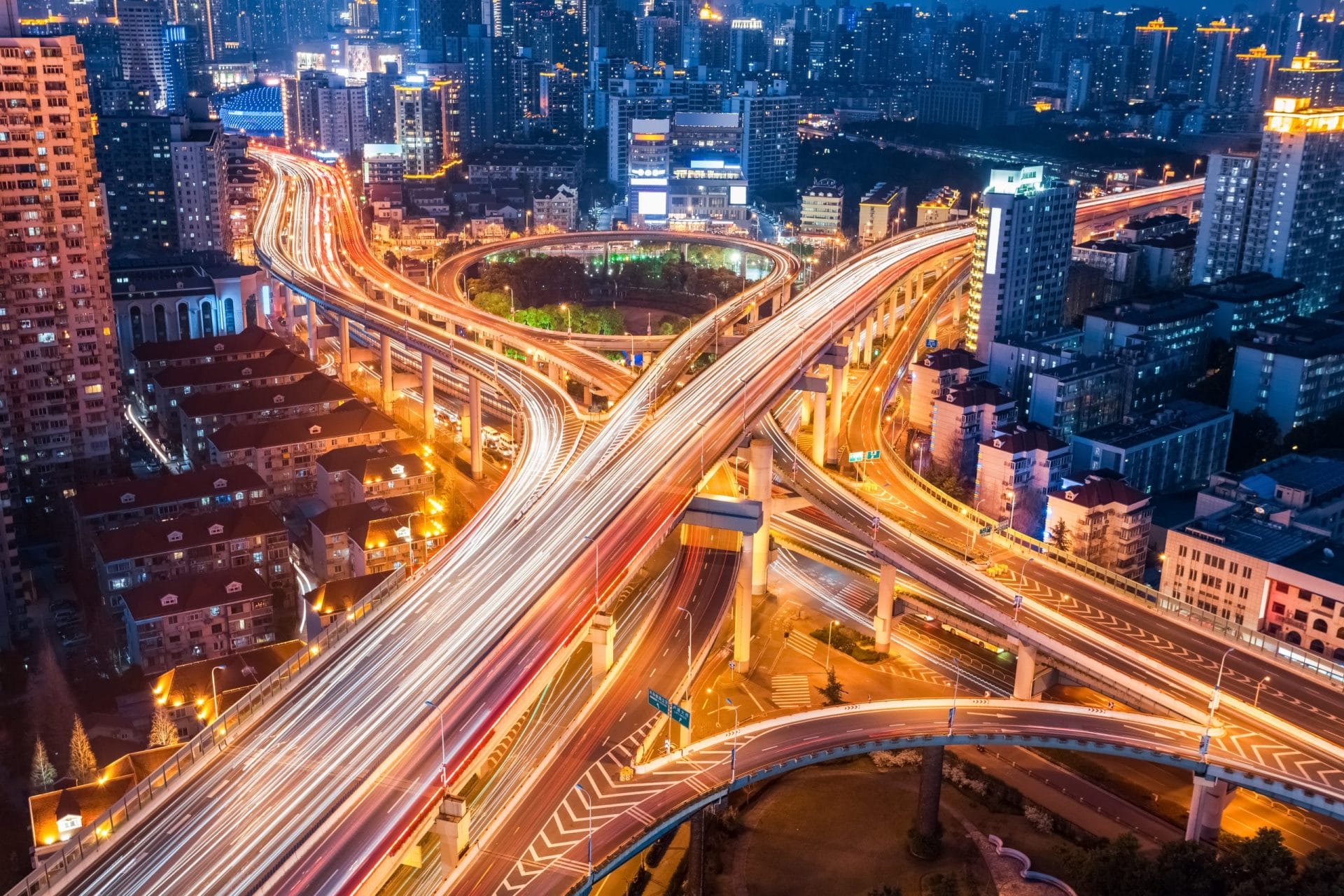 A vibrant cityscape at night shows intertwining highways and overpasses with bright, blurred lights from moving vehicles, surrounded by tall buildings and illuminated by streetlights.