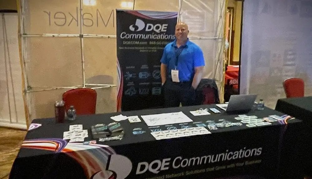 A person stands behind a DQE Communications booth at an event, with promotional materials and branded items displayed on the table and a large company banner in the background.