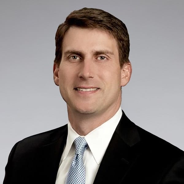 A man in a dark suit, white dress shirt, and light blue patterned tie, smiling against a plain gray background.