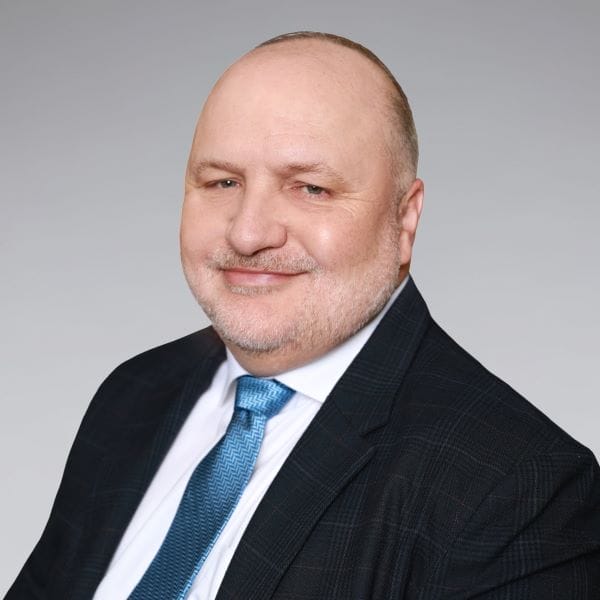 A middle-aged man with a receding hairline and short beard, wearing a dark checkered suit, white shirt, and blue patterned tie, smiles at the camera against a plain gray background.