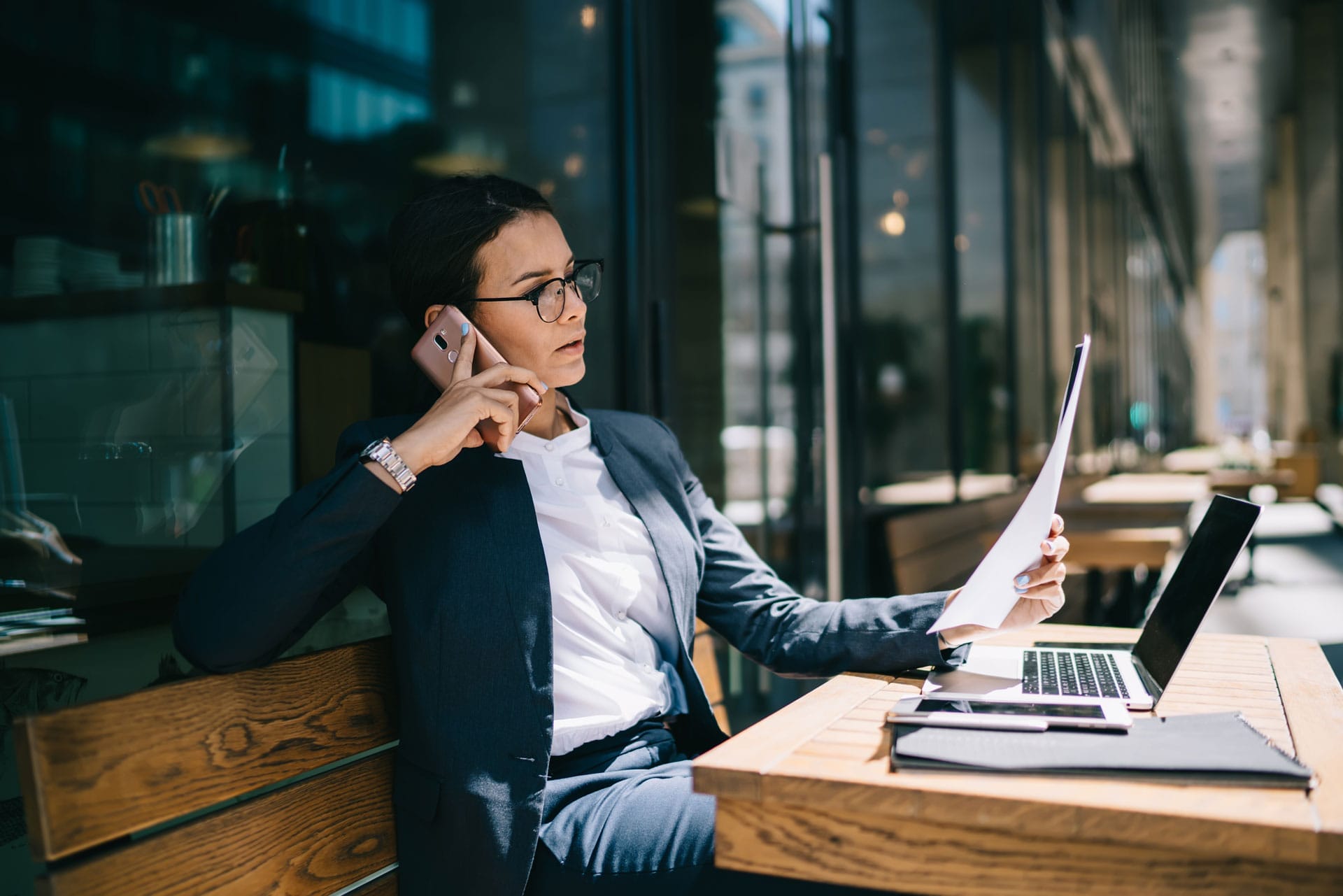 A woman in a suit sits at an outdoor café table, talking on her phone while holding documents. A laptop and notebook are open in front of her. She appears focused and professional.