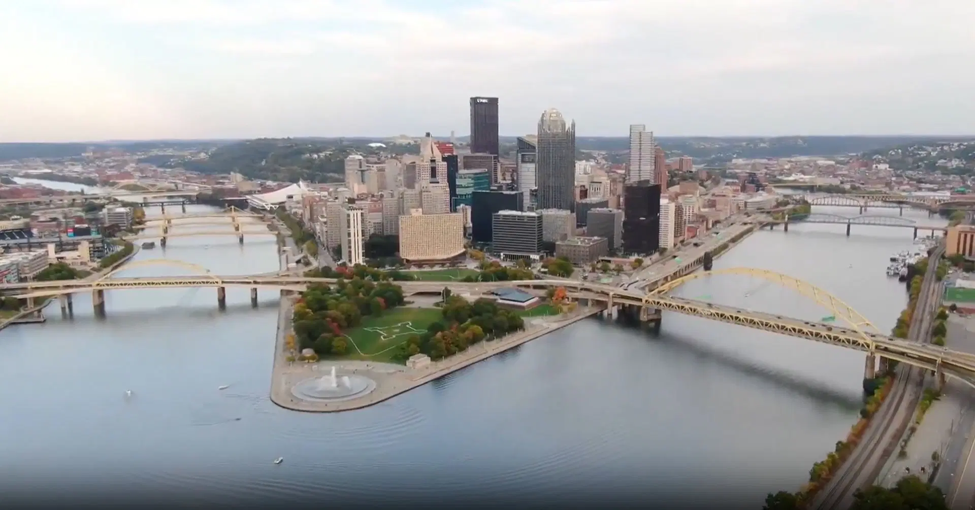 Aerial view of downtown city skyline with tall buildings, rivers converging, yellow bridges, green park areas, and a fountain at the riverfront under a cloudy sky.
