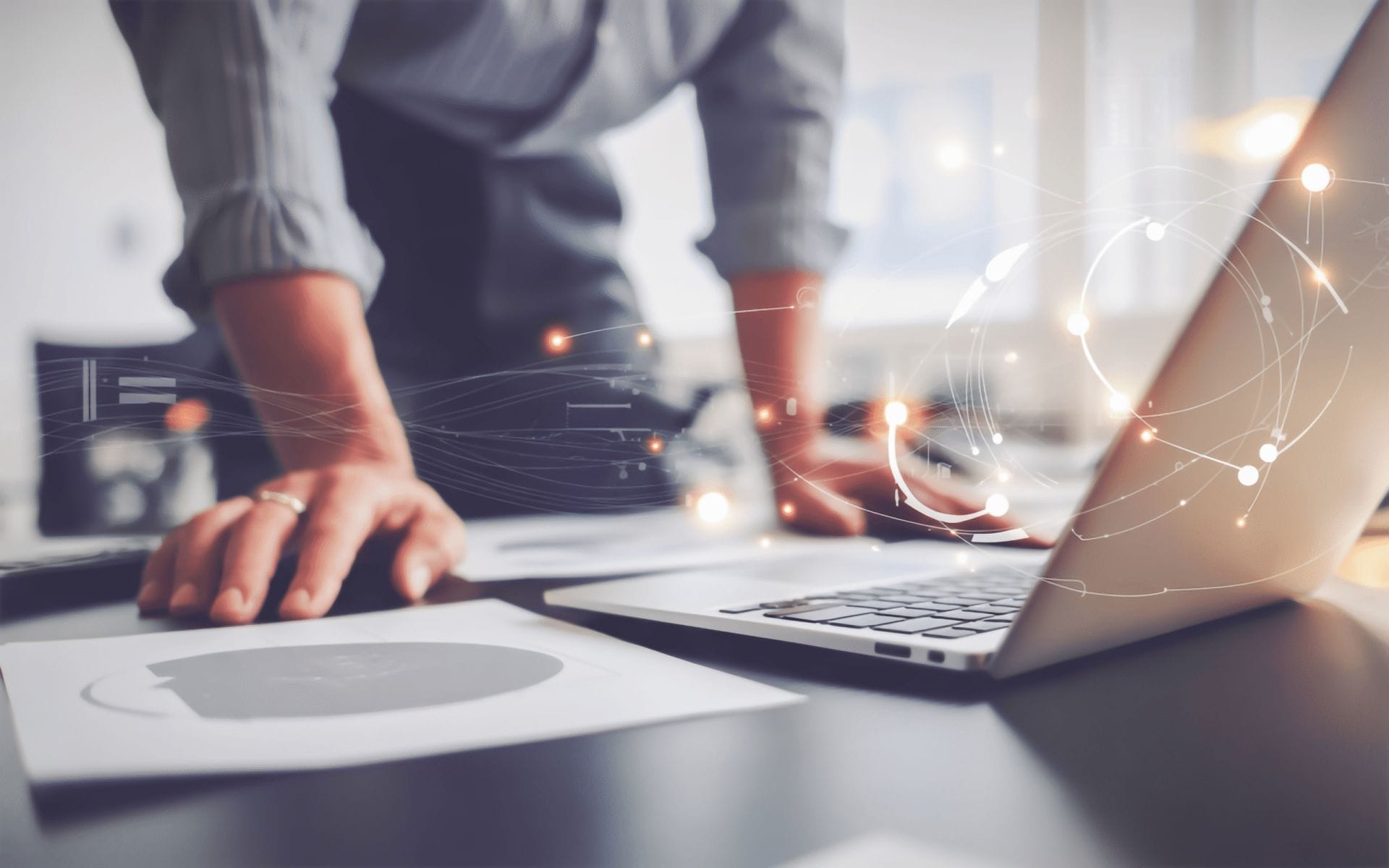 A person stands over a desk, leaning on their hands beside a laptop and some documents. Abstract digital network graphics overlay the scene, suggesting technology or data analysis.