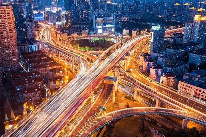 Aerial view of a brightly lit city at night, featuring a complex network of elevated highways with streaks of headlights and taillights, surrounded by tall buildings and city lights.