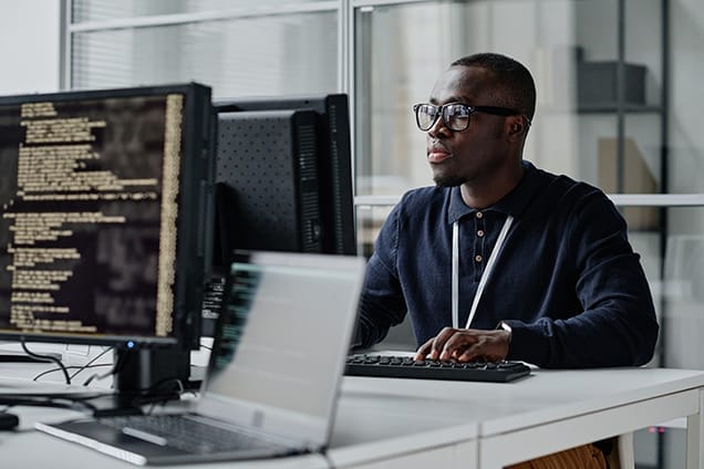 A man wearing glasses types on a keyboard at a desk with two computer monitors displaying code in an office setting. He appears focused on his work.