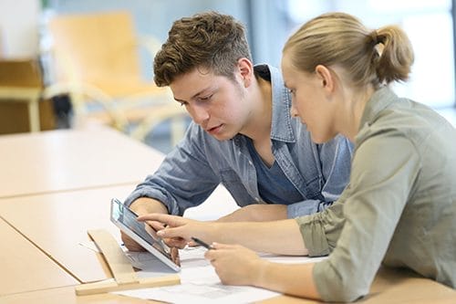 Two young adults sit at a table, looking at a tablet together. One person points at the screen while the other holds a pen, with papers spread out on the table in front of them.