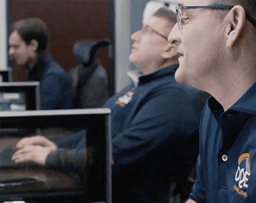 Three people sit at desks in an office setting, focused on computer screens. They are wearing matching navy blue shirts with a yellow and white logo on the chest. The environment appears professional and collaborative.