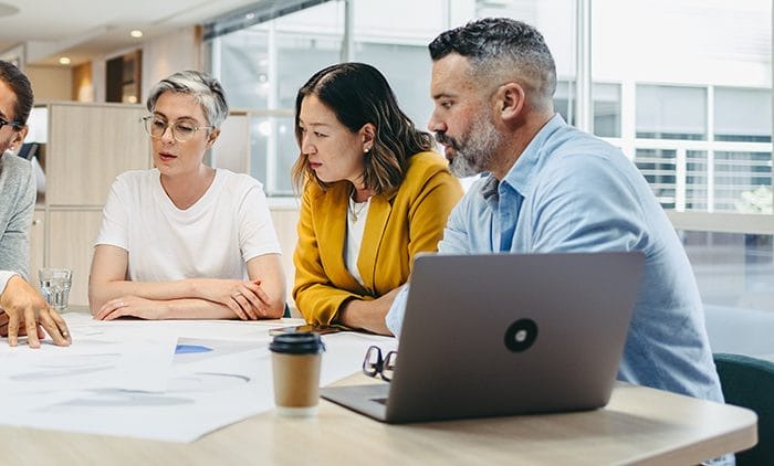 Three people sit at a table in an office, looking at documents and a laptop. They appear to be engaged in a discussion or meeting. A coffee cup and papers are also on the table.