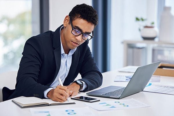 A man in a suit and glasses writes in a notebook at a desk with a laptop, smartphone, and paperwork with charts in a bright office setting.