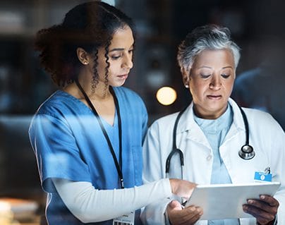 Two healthcare professionals, one in blue scrubs and the other in a white coat with a stethoscope, discuss information displayed on a digital tablet in a medical setting.