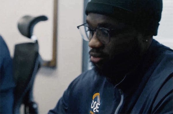 A man wearing glasses, a dark beanie, and a jacket with an "ODE" logo sits indoors, looking down and slightly to the side. There is a blurred office chair in the background.
