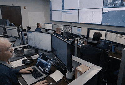 Several people work at computer stations in a control room filled with large monitors displaying data, maps, and surveillance footage. The atmosphere appears focused and high-tech.
