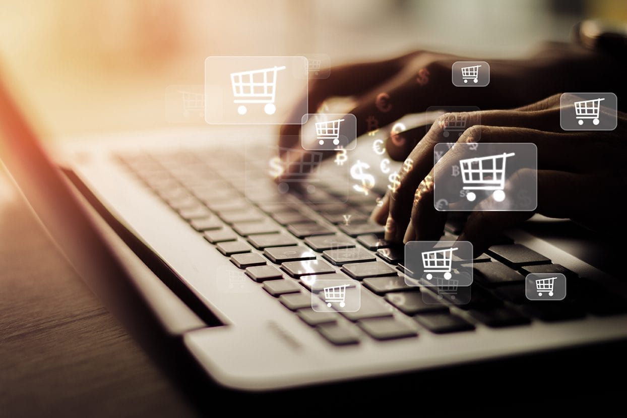 Hands typing on a laptop keyboard with digital shopping cart icons and currency symbols floating above the keys, representing online shopping or e-commerce.
