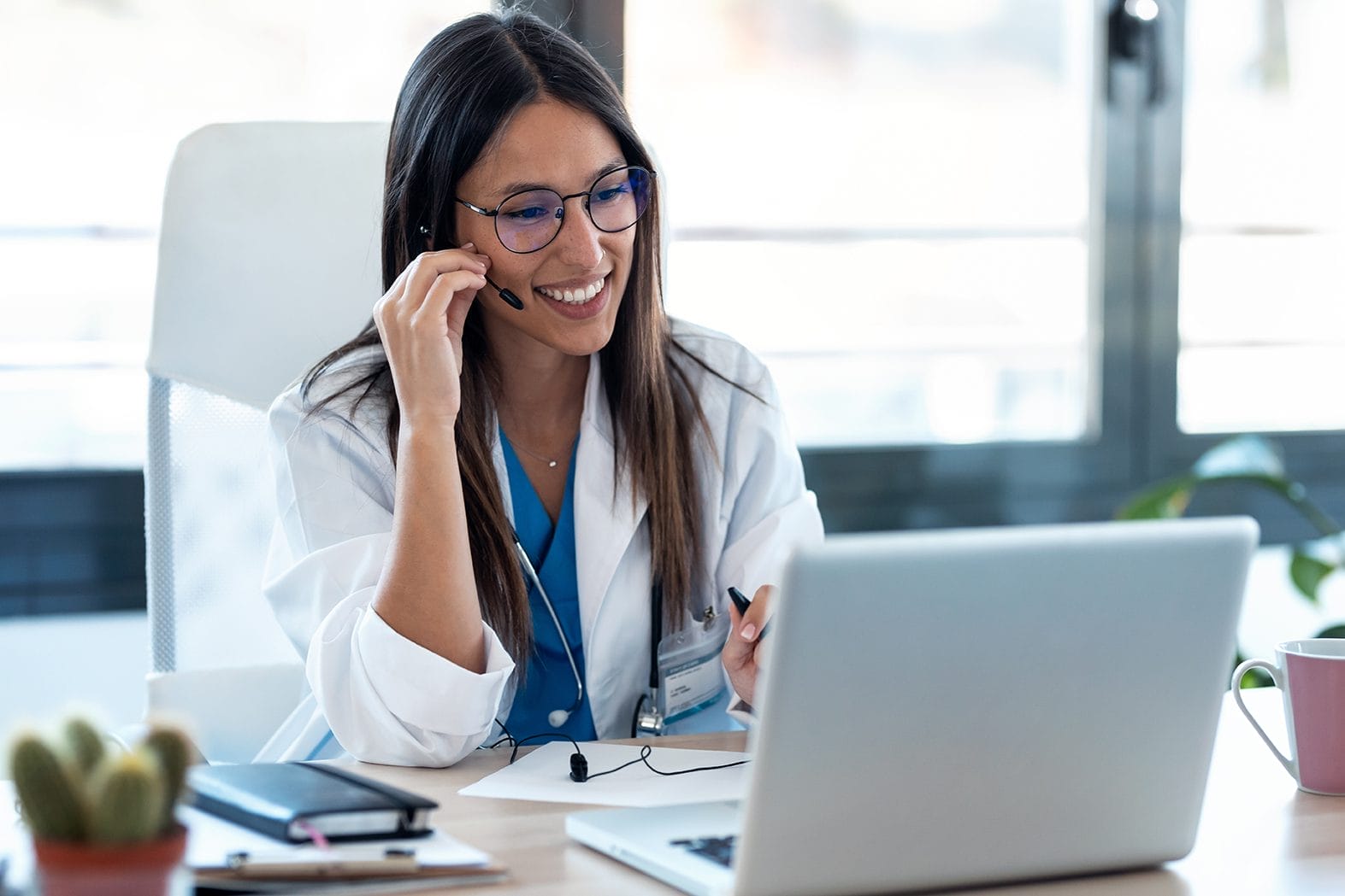 A smiling doctor wearing glasses and a white coat sits at a desk, using a headset and looking at a laptop during a video call. Notebooks and a coffee mug are on the desk in front of her.