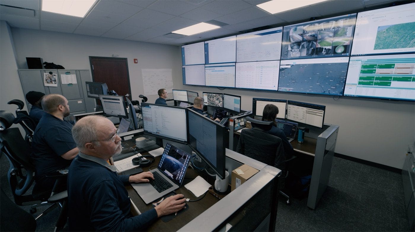 Several people work at computer desks in a control room, monitoring multiple large screens on the wall that display data, maps, and video feeds. The room has office chairs, equipment, and a busy, focused atmosphere.