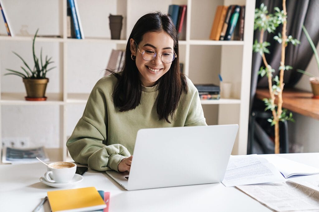 A young woman with glasses and a green sweater smiles while using a laptop at a white desk. There are notebooks, documents, a cup of coffee, and bookshelves with plants in the background.