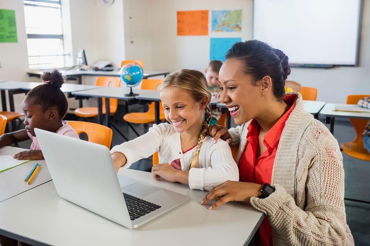 A teacher and a young student sit together at a classroom desk, smiling and looking at a laptop screen. Other students are working at their desks in the background.