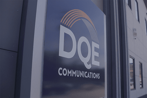 Close-up of a sign on a building displaying the logo and name "DQE Communications" with an orange arc above the letters, reflecting part of the building and sky in the background.