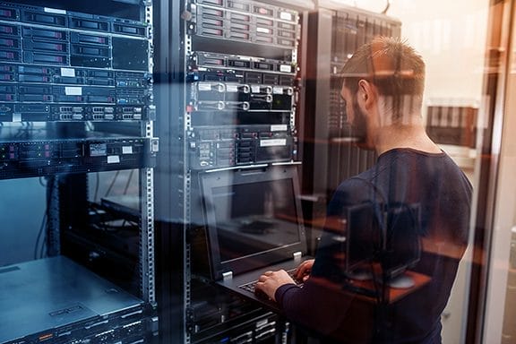 A person is working on a laptop in a server room, surrounded by server racks and networking equipment. The scene shows a professional environment focused on IT infrastructure management.