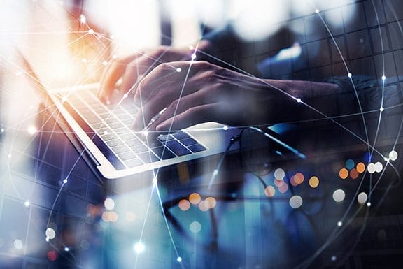 Hands typing on a laptop keyboard with a digital overlay of connected lines and nodes, suggesting technology, networking, or internet connectivity. Blurred lights and reflections are in the background.