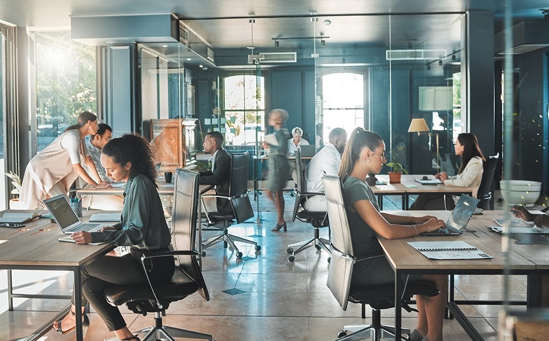 Modern open-plan office with people working at desks on laptops, some collaborating in small groups, others focused individually. Large windows let in natural light, and the workspace has a professional, bright atmosphere.