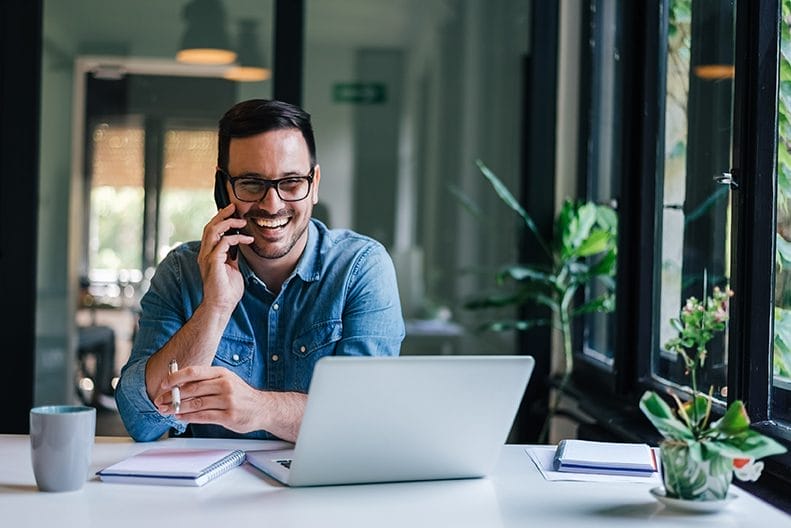 A smiling man wearing glasses and a denim shirt sits at a desk, talking on his smartphone, with an open laptop, notebook, and coffee mug in front of him. Large windows and green plants are visible in the background.