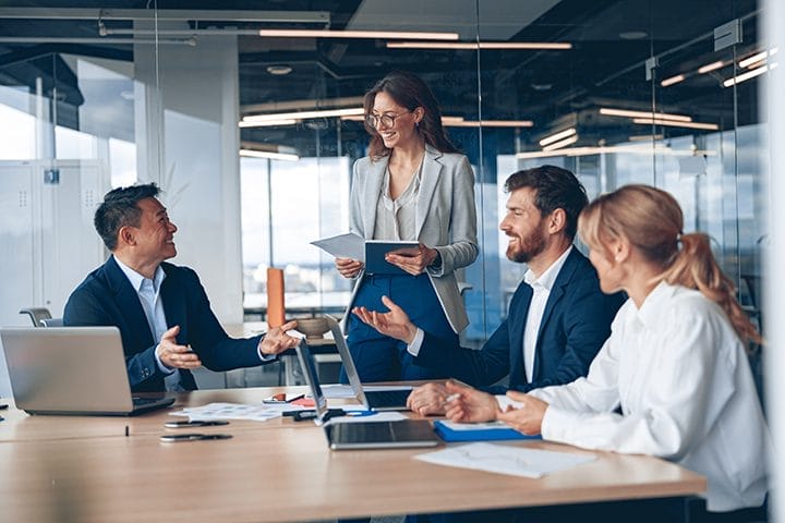 Four business professionals sit at a table in a modern office, discussing work. One woman stands holding documents, smiling, while the others listen. Laptops, papers, and pens are on the table.