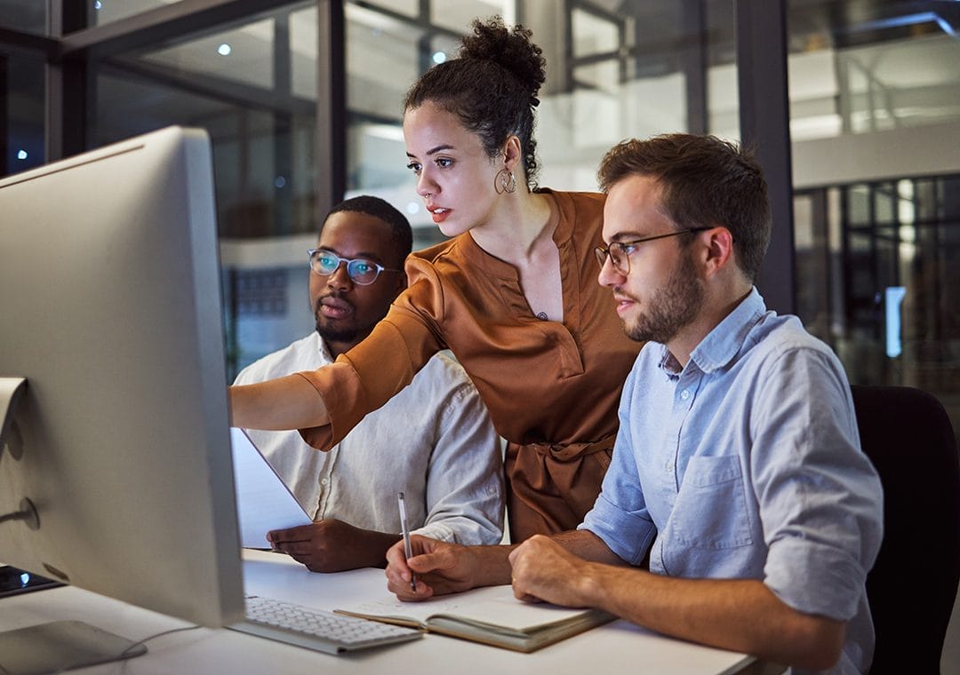 Three colleagues work together at a desk, looking intently at a large computer monitor. One woman is pointing at the screen while two men watch and take notes, suggesting a collaborative office environment.