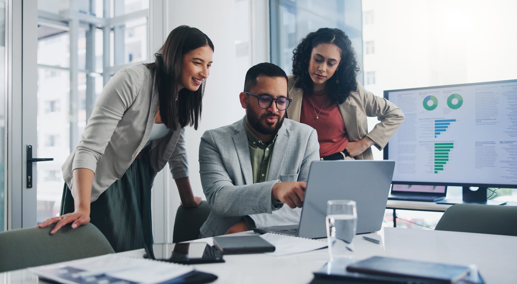 Three colleagues in business attire gather around a laptop in a modern office, reviewing enterprise fiber solutions for Lancaster PA. Charts and graphs are visible on a monitor in the background.